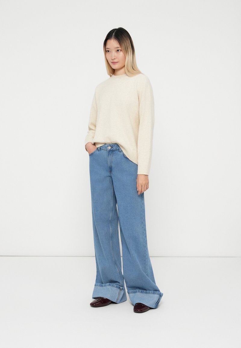 Beżowy sweter dzianinowy z okrągłym dekoltem, w połączeniu z jasnoniebieskimi spodniami o szerokich nogawkach z podwiniętymi mankietami. Modelka stoi w luźnej pozie z ręką w kieszeni.