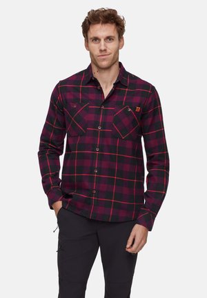 Homme portant une chemise à carreaux rouge et noir avec poches, se tenant contre un fond blanc uni, les mains partiellement dans les poches.