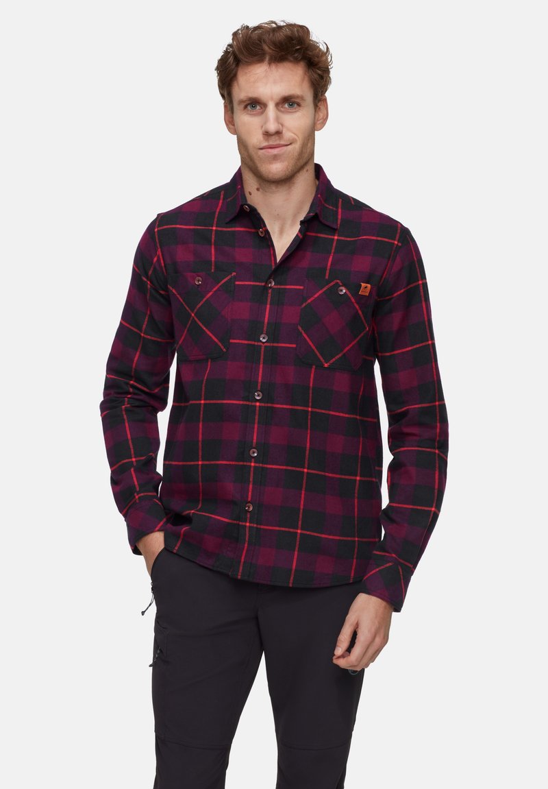 Uomo che indossa una camicia button-up a quadri rossi e neri con tasche, in piedi davanti a uno sfondo bianco semplice, con le mani parzialmente nelle tasche.