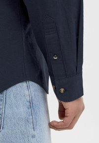 Camicia a maniche lunghe blu navy con polsini abbottonati, caratterizzata da una trama leggera e un orlo curvo, abbinata a jeans azzurri chiari.