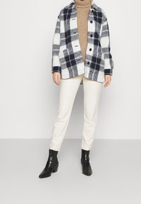 Navy jaalilla ja valkoisella villasekoitteen takki, jossa on napit ja läpätaskut. Yhdistettynä kermaisiin slim-fit-housuihin ja mustiin nilkkasaappaisiin.