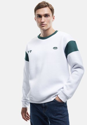 Sudadera blanca con detalles verdes en las mangas y el cuello, presenta un logo y un corte relajado. Combinada con pantalones de mezclilla azul.