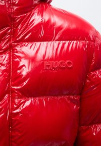 Piros pufi dzseki fényes felülettel, steppelt mintával és dombornyomott „HUGO" logóval. Kapucnival és vízszintes varrási részletekkel rendelkezik.
