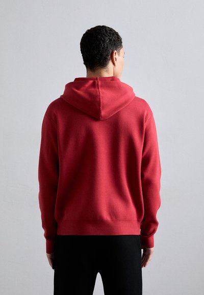 Felpa rossa con tasca a marsupio, polsini e orlo a coste. Tessuto liscio con cappuccio regolabile, indossata con pantaloni neri.