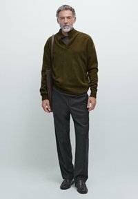 Man met een grijze baard, gekleed in een olijfgroene sweater, donkere broek, zwarte schoenen en met een bruine schoudertas, staande tegen een egale achtergrond.
