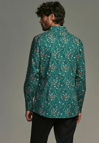 Cămașă verde cu mânecă lungă și model floral, având accente roșii, albastre și albe; guler în stil butonat și tiv curbat. Textură netedă.