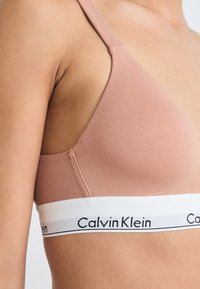 Sujeta sin aros de Calvin Klein en suave tejido rosa con un diseño triangular. Presenta una amplia banda blanca con detalle del logo en negro.