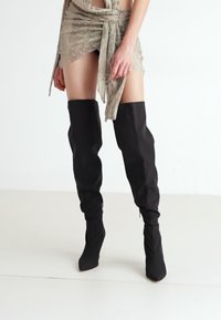 En person iført et kort beige skjørt med knytedetalj foran og svarte knelange spissede støvler står på et hvitt gulv.