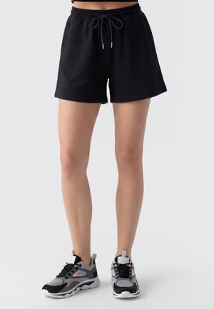Personne portant un short noir à cordon et des baskets de sport noires, grises et blanches, debout sur un fond clair.