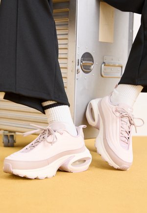 Person iført lyserøde Nike sneakers med hvide sokker og sorte bukser, fødderne hviler nær en metallisk opbevaringskasse på gul gulv.