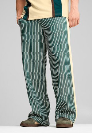 Person, die grüne und weiß vertikal gestreifte Hosen mit gelben Seitenstreifen und grünen Schuhen vor einem grauen Hintergrund trägt.
