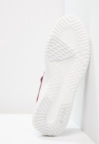 Röd sportskor med texturerad karminöverdel, vit gummisula med mönstrad slitbana för grepp, lutad mot en vit bakgrund.