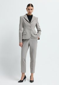 Femme portant un blouson cintré gris clair et un pantalon assorti avec des talons noirs texturés, posant sur un fond blanc uni.