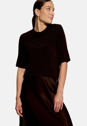 Femme portant un pull noir à manches courtes et une jupe noire, regardant vers sa droite, cheveux attachés et portant de petites boucles d’oreilles créoles.