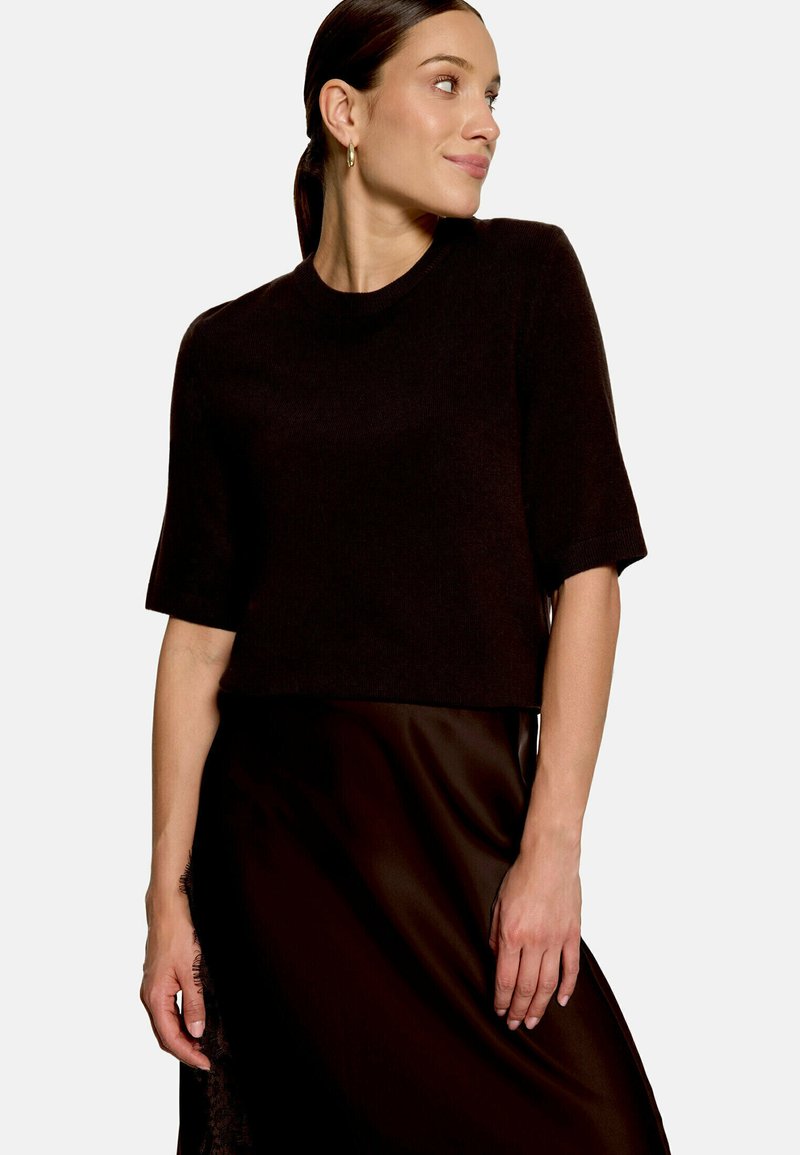 Femme portant un pull noir à manches courtes et une jupe noire, regardant vers sa droite, cheveux attachés et portant de petites boucles d’oreilles créoles.