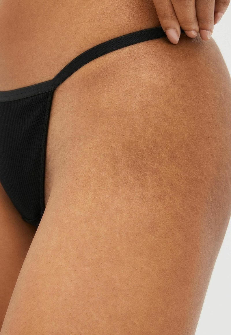 Slip di bikini neri con spalline sottili, texture a coste e design minimale. Pelle con evidenti smagliature nell'area attorno alla coscia.