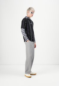 Ung person med kort platinahår, iført hvide solbriller, en lagdelt sort t-shirt over en ternet langærmet skjorte, grå bukser og hvide sneakers.