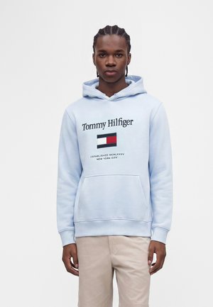 Jonge man met gevlochten haar, dragend een lichtblauwe Tommy Hilfiger hoodie en beige broek, staand tegen een effen witte achtergrond.