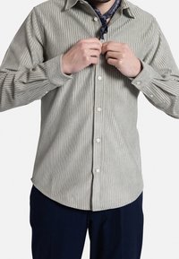 Camisa de botones a rayas verde claro, hecha de una tela texturizada, con cuello y mangas largas, parcialmente abotonada y una bandana azul marino visible.