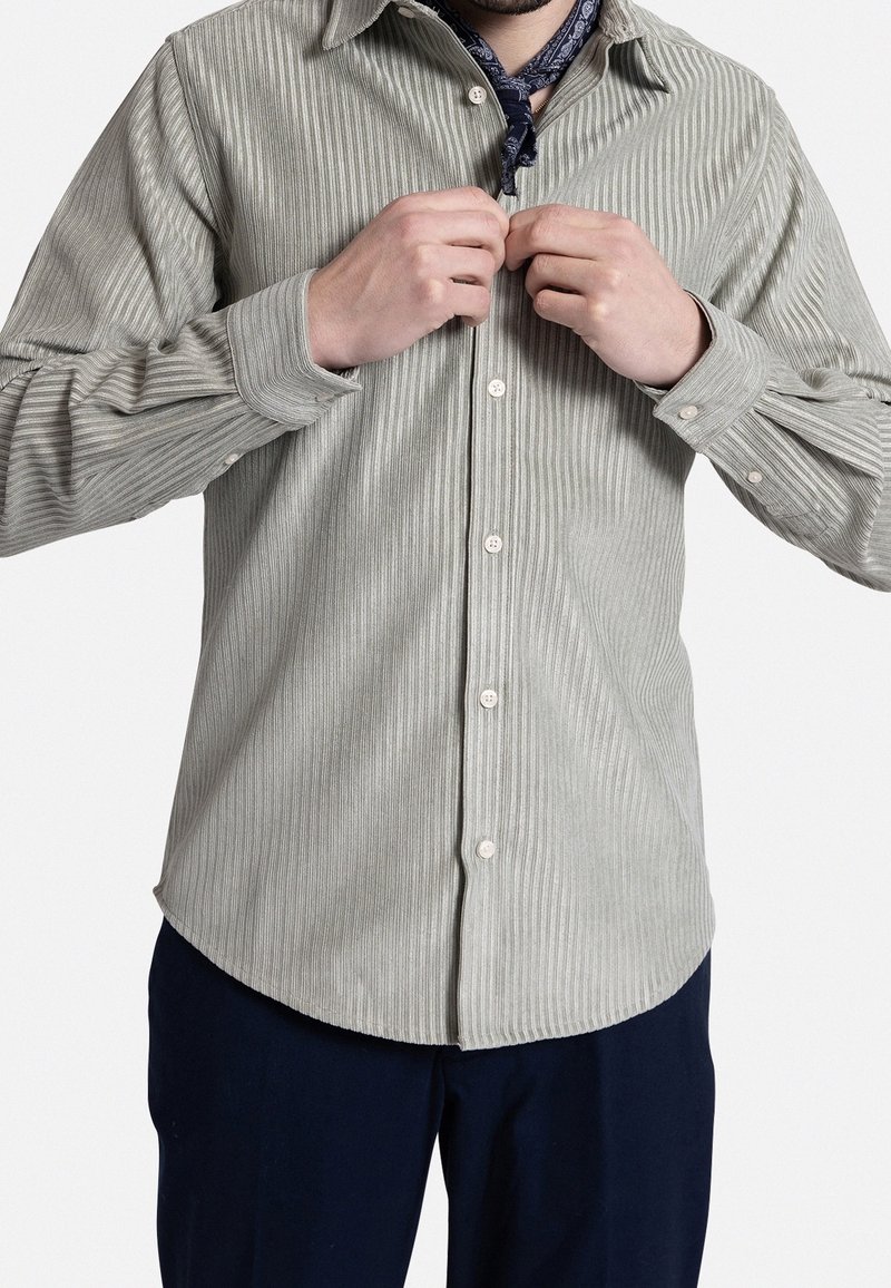 Camisa de botones a rayas verde claro, hecha de una tela texturizada, con cuello y mangas largas, parcialmente abotonada y una bandana azul marino visible.