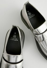 Zapatos deslizables metálicos plateados con suela plana, que cuentan con dos correas en la parte superior y un acabado suave y brillante. Forro interior negro.