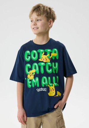 Chłopiec z blond włosami nosi granatową koszulkę Pokémon z zielonym napisem „Gotta Catch 'Em All!” i trzema postaciami Pikachu, stoi z ręką w kieszeni.