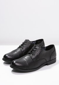 Chaussures habillées en cuir noir avec une texture lisse, un design à bout golf, des détails perforés et un bout arrondi. Dotées d'une semelle noire robuste.