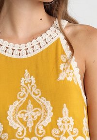 Keltainen kirjailtu paita, jossa on virkattu kaula-aukko, monimutkaiset valkoiset kukkakuviot ja pehmeä kangas.