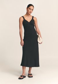 Črna midi obleka z nagubanimi naramnicami, zavitim zgornjim delom in oprijeto silhueto. Oblečena s ploskimi črnimi sandali in drži kremasto teksturirano torbico.