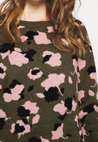 Donna che indossa un maglione verde oliva con un grande motivo floreale astratto in rosa e nero, visibile dal petto al collo, con capelli ondulati di colore marrone chiaro.