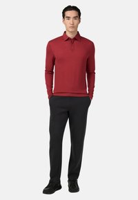 Mladý muž stojící s rukama v kapsách černých kalhot, oblečený v červeném polo tričku s dlouhým rukávem a černých teniskách na bílém pozadí.