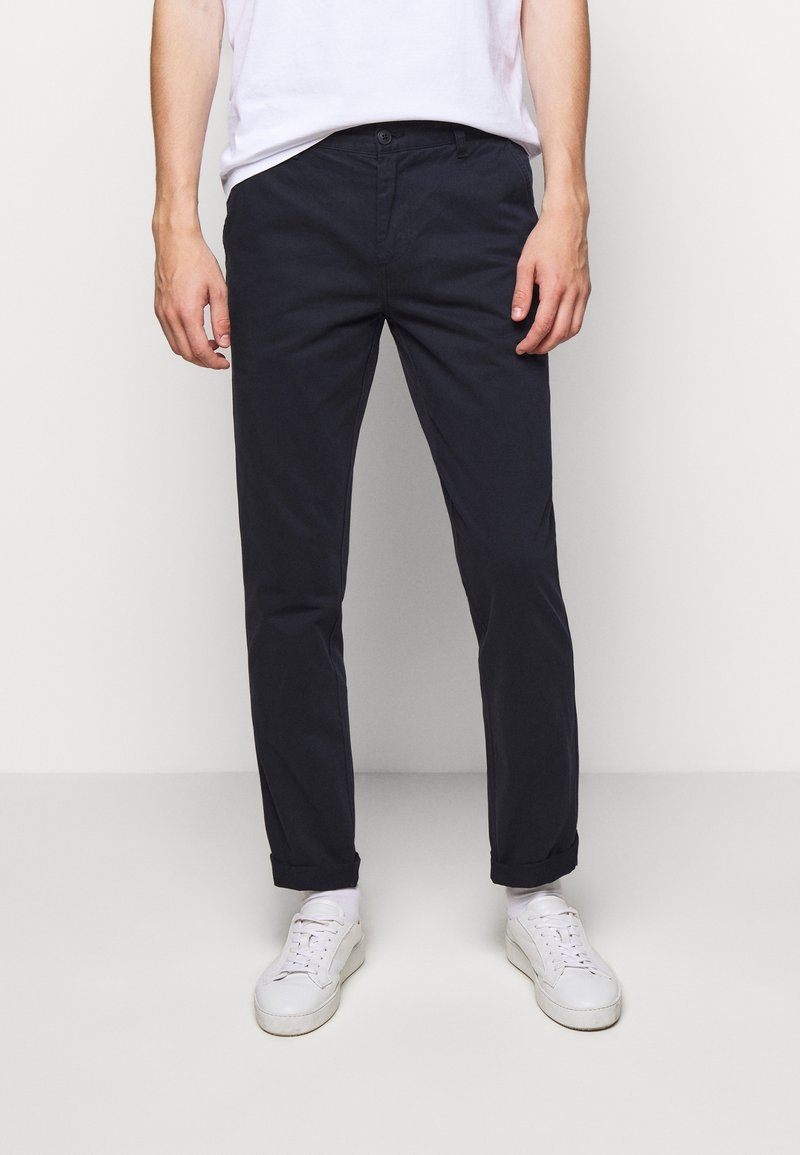 Marineblauwe katoenen chinos met een rechte pasvorm, zijzakken en een knoopsluiting aan de voorkant, gedragen met witte sneakers en lichte sokken.