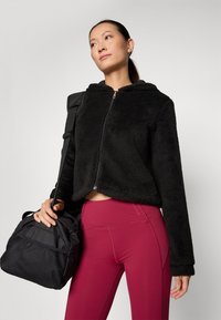 Giacca in pelliccia sintetica nera con zip e cappuccio, caratterizzata da un design corto. Abbinata a leggins aderenti bordeaux e a una borsa a secchiello nera.