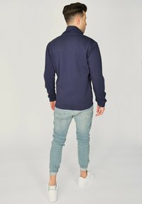 Granatowa polarowa kurtka na zamek z wysokim kołnierzem, gładką fakturą i dopasowanymi mankietami, zestawiona z jasnoniebieskimi slim fit dżinsami i białymi trampkami.