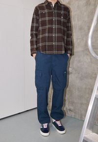 Chemise à carreaux marron avec des accents verts, pantalon cargo bleu marine avec poches et baskets multicolores avec une semelle blanche.