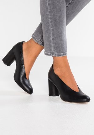 Zapatos de tacón alto de cuero negro con punta redondeada y un diseño distintivo con recortes, que cuenta con una superficie texturizada y un tacón grueso.