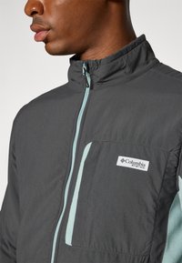 Chaqueta gris con cuello alto y cremallera azul claro. Presenta un bolsillo en el pecho con cremallera y un logo de Columbia en el bolsillo. Textura suave.