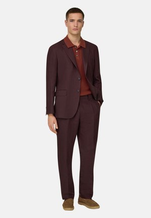 Ung mand iført en mørkebrun jakkesæt med en rustfarvet strikket polo og lysebrune ruskindssko, stående med den ene hånd i lommen.