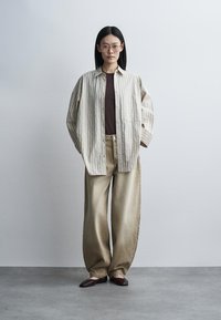 Femme avec des lunettes debout portant une chemise beige à rayures boutonnée sur un haut marron foncé, un pantalon beige ample et des ballerines marron devant un fond uni.