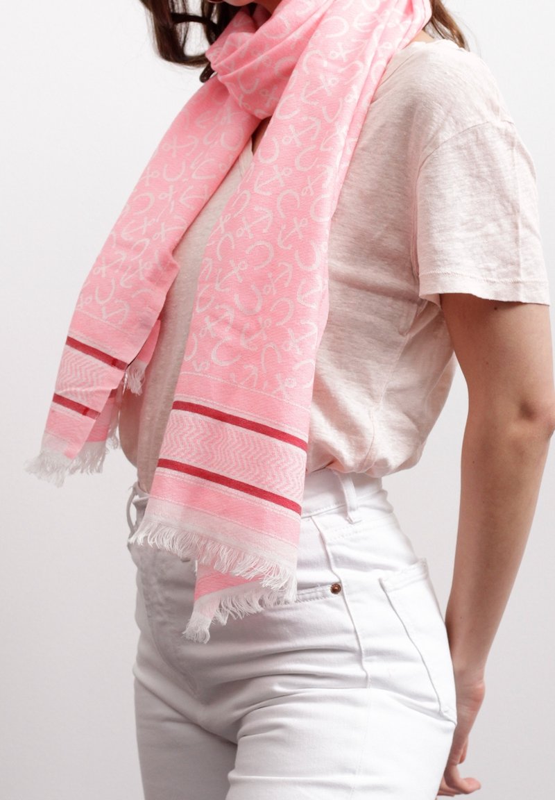 Écharpe rose avec des ancres blanches et des rayures rouges portée sur un t-shirt beige et un pantalon blanc sur une personne debout.