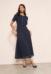 Tengerészkék pliszírozott ruha rövid ujjakkal és gombokkal, struktúrált felsőrésszel és áramló szoknyával, lila magassarkú cipővel párosítva.