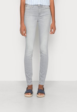 Persona che indossa jeans aderenti grigio chiaro e sandali aperti blu scuro, in piedi contro uno sfondo chiaro semplice.