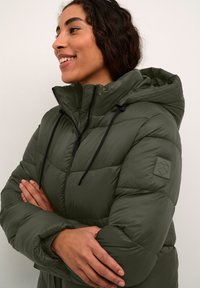 Blouson matelassé vert olive avec capuche, présentant une fermeture éclair à l'avant, un cordon de serrage réglable et un motif texturé sur les manches.