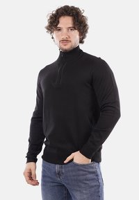 Maglione nero lavorato a maglia con colletto alto e chiusura a mezza zip. Presenta maniche lunghe e un design aderente. Abbinato a jeans blu.