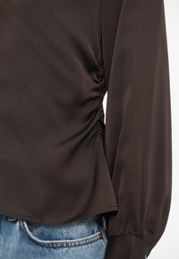 Kitsa mustriga tumepruun pikka varrukaga pluus, millel on kokku volditud külgedetail, kantud heleda sinise teksapükstega.