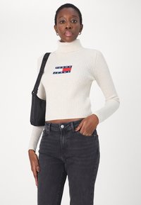Krémový pletený rolák s barevným logem "TOMMY JEANS" na přední straně, spárovaný s tmavě šedými džíny a černou kabelkou přes rameno.