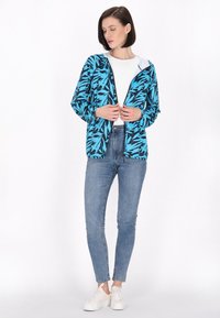 Giacca leggera con zip di un vivace blu, decorata con un motivo di foglie nere, abbinata a una maglietta bianca e a jeans skinny blu. La texture appare liscia.