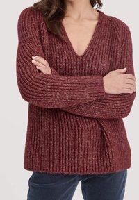 Femme portant un pull en maille côtelée rouge profond à col en V et manches longues, les bras croisés, assorti à un pantalon en velours côtelé bleu foncé.