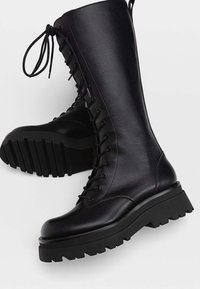 Botas de cuero negro hasta la rodilla con suelas gruesas y cordones frontales, mostrando una bota en posición vertical y la otra de lado.