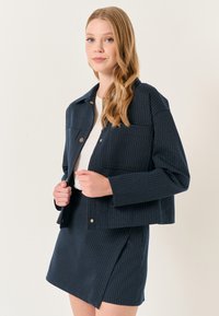 Marineblauwe pinstripe blazer met gouden accenten, met een boxy ontwerp en twee voorzakken, gecombineerd met een bijbehorende rok.